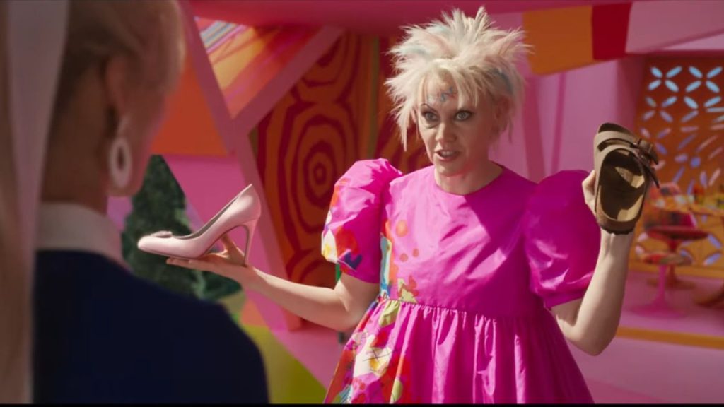 A scene from the Barbie movie where a character with spiky, wild hair in a bright pink, puff-sleeved dress is holding up two shoes: a high heel in one hand and a Birkenstock sandal in the other. The background features a colorful, vibrant setting. 